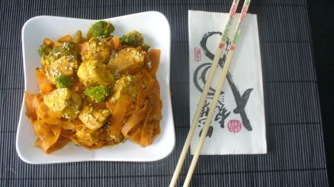 Eine asiatische Möhrengemüsepfanne mit Hähnchenbrust. Eine farbenfrohe Möhrengemüsepfanne mit Hähnchenbrust liegt in einer weißen Schüssel, umgeben von Essstäbchen und einer Serviette.