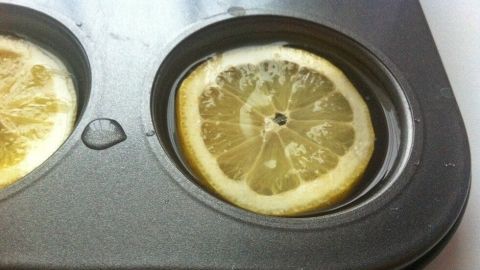 Ein Muffinblech zeigt große Eiswürfel in der Form mit einer ganzen Zitronenscheibe darin, umgeben von Wasser.