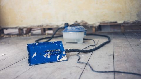 Ein blauer Styroporkleber-Entferner liegt auf dem Boden, während er eine Wand in einem renovierungsbedürftigen Raum bearbeitet.