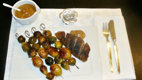 Gegrillte Rosenkohl-Kartoffel-Spieße liegen neben gebratener Cranberry-Entenbrust auf einem eleganten Teller, umgeben von Soße.