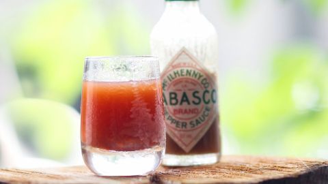 Ein Glas mit einer scharfen, roten Flüssigkeit steht neben einer Flasche Tabasco-Sauce auf einem Holzuntergrund.