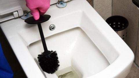 Ein Putzgerät schwappt im WC, während eine Hand mit pinker Gummihandschuh einen Urinstein entfernt. Toilettenumfeld sichtbar.