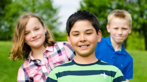 Drei Kinder lächeln fröhlich im Freien, stehen auf einer grünen Wiese und genießen die gemeinsame Spielzeit draußen.