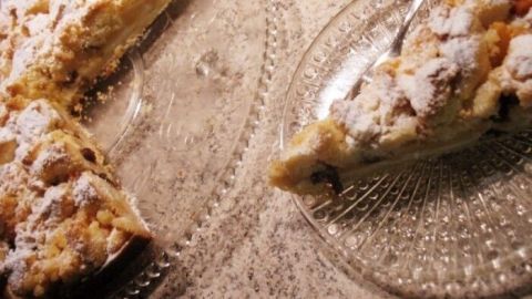 Eine Krümeltorte, bestäubt mit Puderzucker, wird auf einem Glasplatte präsentiert, während ein Stück auf einem Teller serviert wird.