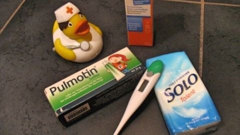 Eine Gummiente mit Krankenpflegerhut liegt neben Medikamenten, einem Fieberthermometer und einer Verpackung für Lutschtabletten.