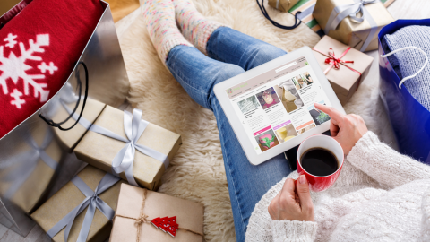 Eine Person sitzt auf einem Teppich, blickt auf ein Tablet und genießt einen warmen Tee, umgeben von Weihnachtsgeschenken.