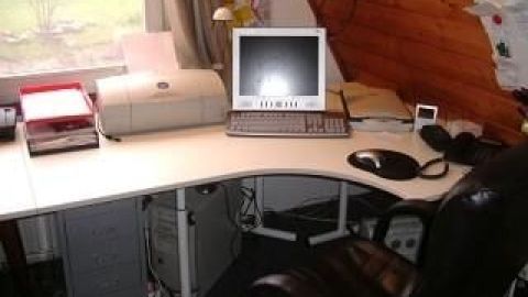 Ein weißer IKEA-Bürotisch steht in einem Dachzimmer, bestückt mit Monitor, Drucker und Schreibtischsessel; Fenster daneben.