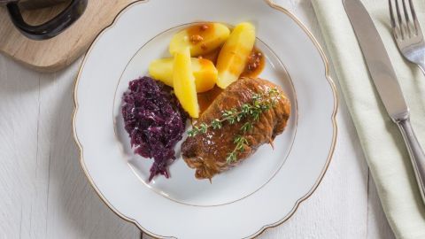 Saftige Wildschweinrouladen liegen auf einem weißen Teller, begleitet von Rotkohl und Kartoffeln, in einem rustikalen Setting.