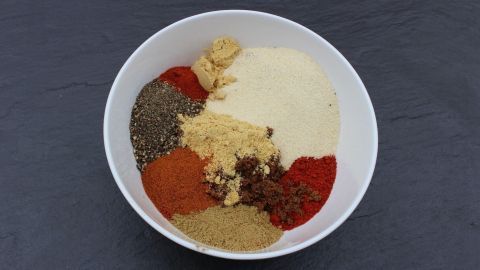 Eine Schüssel mit einer bunten Gewürzmischung liegt auf einem dunkelgrauen Untergrund. Die Gewürze sind in verschiedenen Farben und Texturen angeordnet.
