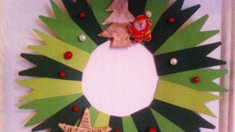 Ein kräftiger grüner Türkranz hängt an einem weißen Hintergrund. Er zeigt weihnachtliche Dekorationen wie einen kleinen Baum und eine Sternform.