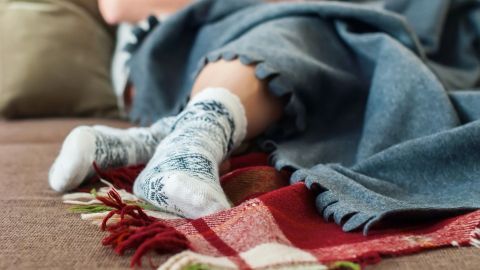 Gemusterte Socken liegen auf einem bunten Teppich, während eine Person gemütlich mit einer Decke drapiert auf einem Sofa sitzt.