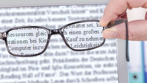 Eine Hand hält eine Brille, die auf einen Bildschirm fokussiert ist, auf dem Beispieltexte für Augenmuskeltraining angezeigt werden.