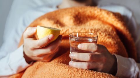 Eine Person sitzt unter einer orangefarbenen Decke, hält eine Zitrone in der einen Hand und ein Glas Wasser in der anderen, um sich gegen die Erkältung abzuhärten.