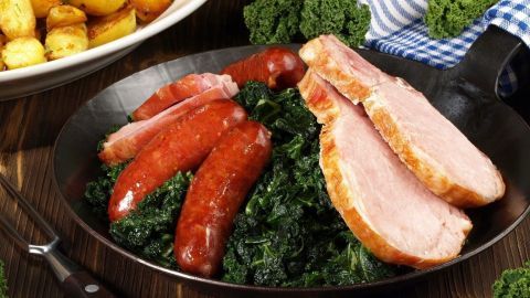 Ein Topf mit Omas ostfriesischem Grünkohl zeigt neben Würstchen und Schinken apetitanlt zu einer herzhaften Mahlzeit, umgeben von Beilagen.