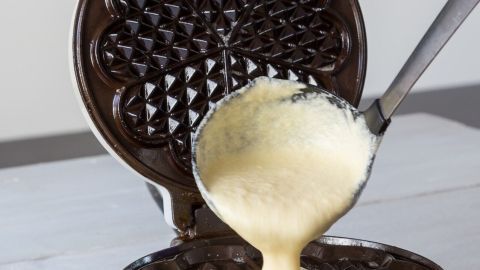 Du möchtest Waffelteig einfrieren und wiederverwenden, weißt aber nicht ob das funktioniert? Waffelteig wird mit einem Löffel in ein Waffeleisen gegossen, das auf einem Holzbrett steht.