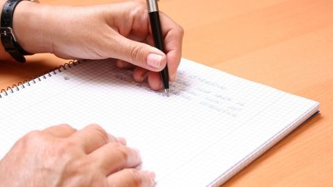 Eine Hand hält einen Stift und schreibt auf ein Notizblatt, während der Tisch eine warme Holzoberfläche zeigt.