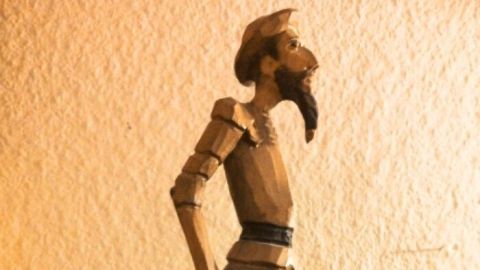 Eine hölzerne Figur steht auf einem Tisch, als würde sie nachdenklich in die Ferne blicken, umgeben von einer neutralen Wand.