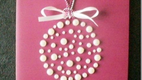 Eine festliche Weihnachtskarte mit Perlen ist auf einem pinken Hintergrund platziert und zeigt eine elegante, perlenbesetzte Dekoration.