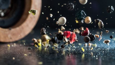 Eine Pfeffermühle kippt, während bunte Pfefferkörner in alle Richtungen verstreut werden, auf einer dunklen Oberfläche.