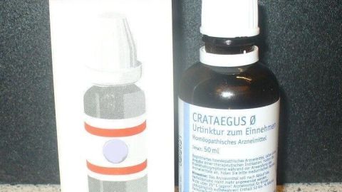 Eine schwarze Flasche mit Crataegus Urtinktur steht neben einer schlichten Verpackung auf einer grauen Oberfläche.