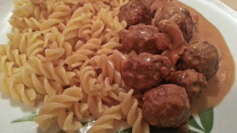 Saftige Köttbullar in Rahmsauce liegen neben spiralförmiger Pasta auf einem Teller, umgeben von einem schlichten Tisch.