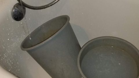 Zwei graue Eimer stehen in einer Badewanne, während Wasser aus einem Wasserhahn hineinläuft, um Wasser zu sparen.