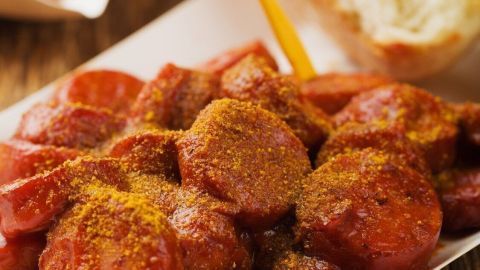 Saftige Currywurst mit Gewürz bestreut, serviert auf einem weißen Teller, umgeben von Pommes und frischem Brot.