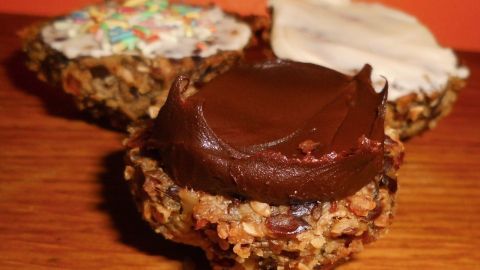 Haferflocken-Leinsamen-Brötchen liegen appetitlich auf einem Holzbrett, zwei sind mit Zuckerguss dekoriert, einer mit Schokoladencreme.