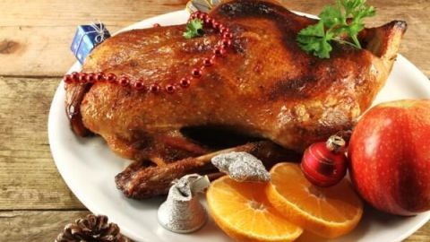 Auf einem weißen Teller liegt eine Weihnachtsente, dekoriert mit einer roten Perlenkette und umgeben von Orangen und Äpfeln.