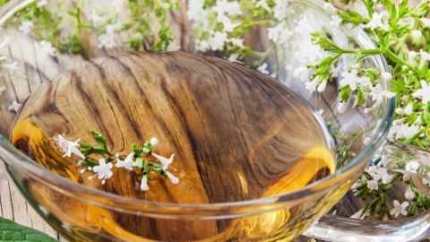 Eine Glas-Schale mit Kräutertee steht auf einem Holztisch, umgeben von frischen Blüten und grünen Blättern.
