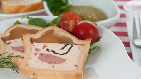 Sliced Terrine liegt auf einem Teller mit Tomaten und Beilage, umgeben von frischem Grün und einer Schüssel Sauce.