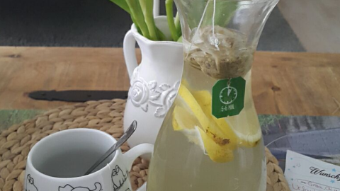Ein Glas mit grünem Tee, frischen Zitronenscheiben und Ingwer steht auf einem Tisch, umgeben von bunten Tulpen.