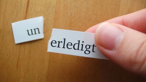 Eine Hand hält ein Papierstück mit dem Wort "erledigt", während ein weiteres Stück mit "un" daneben liegt. Der Hintergrund ist eine helle Holzoberfläche.
