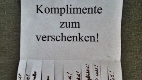 Eine gefaltete Papiernotiz zeigt die Überschrift "Komplimente zum verschenken!" mit mehreren abgetrennten Komplimenten darunter.