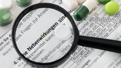 Weißtabletten und Kapseln liegen neben einem Dokument, auf dem "Nebenwirkungen" unter einer Lupe deutlich zu erkennen sind.