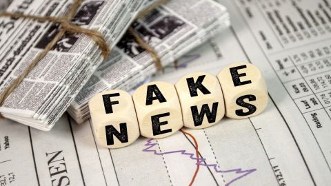 Auf einer Zeitungsoberfläche liegen Holzblöcke mit der Aufschrift "FAKE NEWS". Daneben sind Zeitungen und Datenblätter sichtbar.
