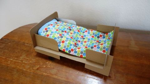 Ein Puppenbett aus Pappe steht auf einem Tisch. Es ist mit einer bunten Decke ausgestattet und bietet eine gemütliche Schlafgelegenheit.