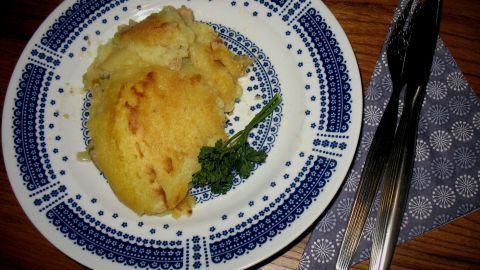 Ein frisch gebackener Hackfleisch-Auflauf mit goldener Kartoffelkruste liegt auf einem blau-musterierten Teller, umgeben von Besteck.