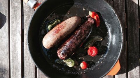 Blut- und Leberwürste braten in einer Pfanne, umgeben von frischen Gemüse auf einer rustikalen Holzoberfläche.