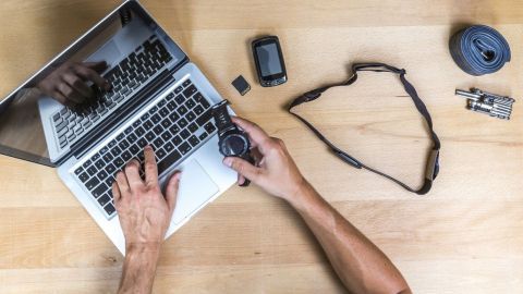 Eine Hand hält ein Trackingsgerät, während sie an einem Laptop arbeitet; daneben liegen weitere Geräte auf einem Tisch.