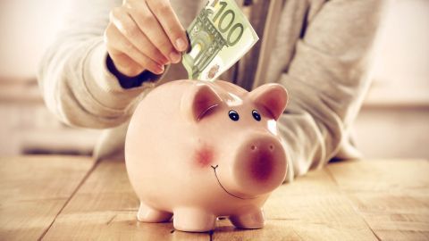 Eine Hand steckt einen 100-Euro-Schein in ein Sparschwein auf einem Holztisch, das für Sparbuch Zinsen steht.