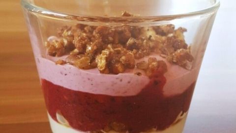 Ein Glas Dessert mit Schichten aus Früchten, Frischkäse und Joghurt, bedeckt mit knusprigem Topping auf einem Holztisch.
