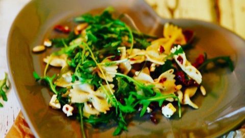 Ein herzförmiger Teller mit Nudelsalat, Rucola und Pinienkernen steht auf einem rustikalen Holztablett, umgeben von dekorativen Salzstreuern.