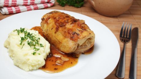 Wirsingroulade liegt auf einem Teller mit Kartoffelbrei und Soße, umgeben von einer roten Tischdecke und einem Topf.