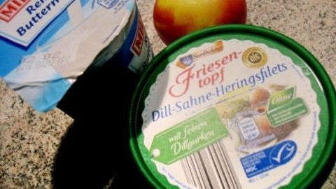 Eine Dose mit Sahnehering und ein Apfel liegen neben einer Packung Buttermilch auf einer grauen Oberfläche.