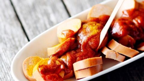 Eine appetitlich angerichtete Currywurst ist mit Sauce überzogen und liegt auf einem weißen Teller, umgeben von Holz.
