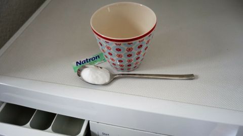 Eine Schale mit roten Punkten steht auf einer weißen Oberfläche neben einem Löffel, während ein Päckchen Natron darauf liegt.