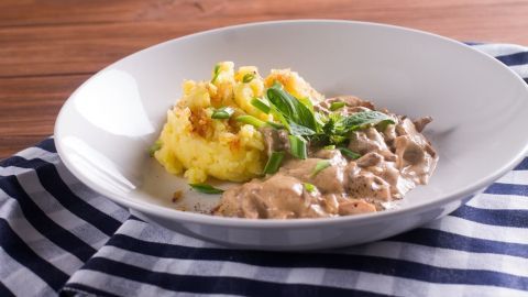 Ein zartes Kalbsleber-Rahmgulasch liegt neben cremigem, gelbem Kartoffelpüree auf einem weißen Teller, alles auf einem gestreiften Tuch.