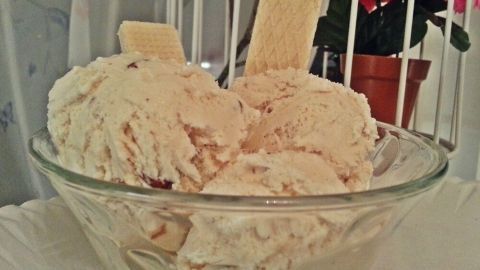 Eine Glasschüssel enthält cremiges Apfel-Zimt-Eis, garniert mit Waffeln, vor einer blühenden Pflanze.