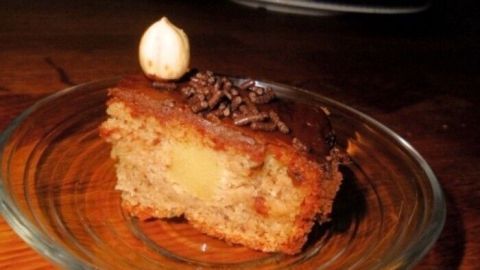 Ein Stück Nusstorte liegt auf einem Glasuntersetzer, während im Hintergrund eine dreistöckige Torte sichtbar ist.